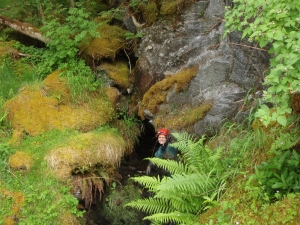 A woman in a red hard hat looks up from the entrance of a cave. The cave is surrounded by lush ferns and moss.