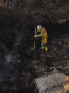 A man in a yellow waterproof suit and a hard hat stands in a cave. He is in knee-deep water and is sampling the water using a long handled net.