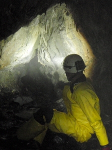 A man in a yellow waterproof suit and hard hat is sat on the ground. He is shining a torch into a limestone cave.